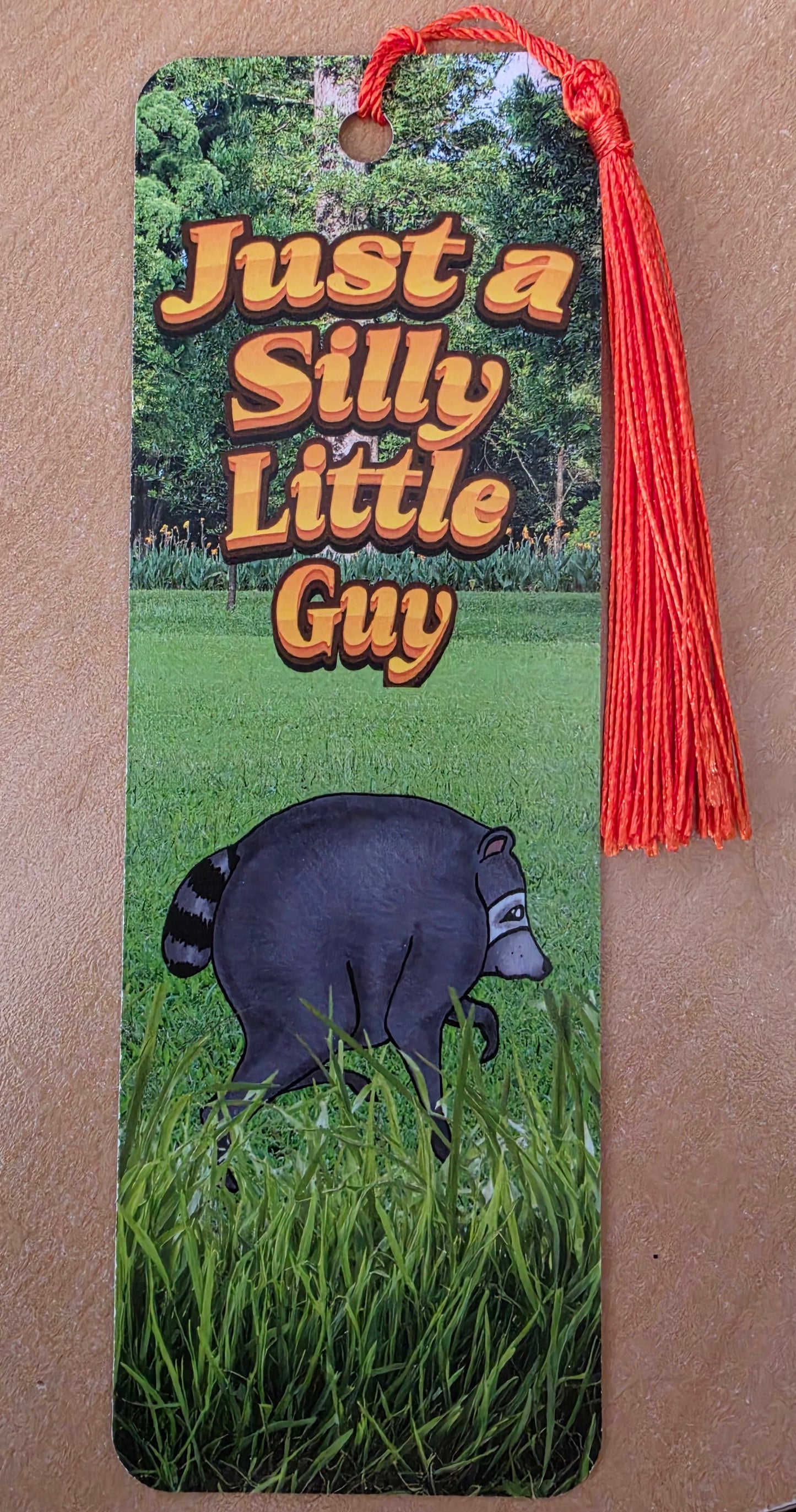 Jimothy Silly Little Guy bookmark with tassel, featuring a cartoon raccoon on a grassy field with bold retro text reading 'Just a Silly Little Guy'